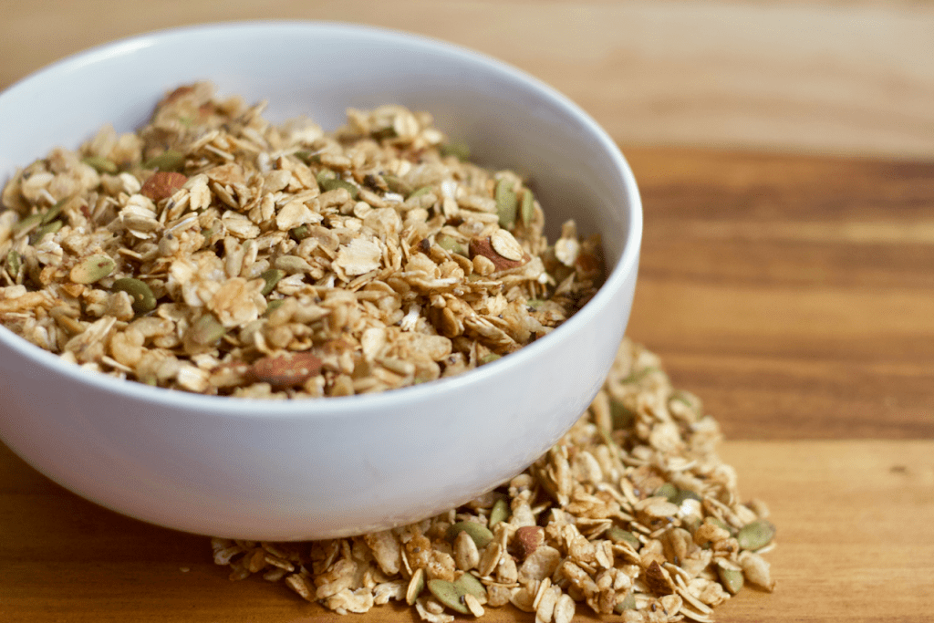 Side view of a bowl full of homemade granola with some on the side of the bowl as well. All on a wooden background.