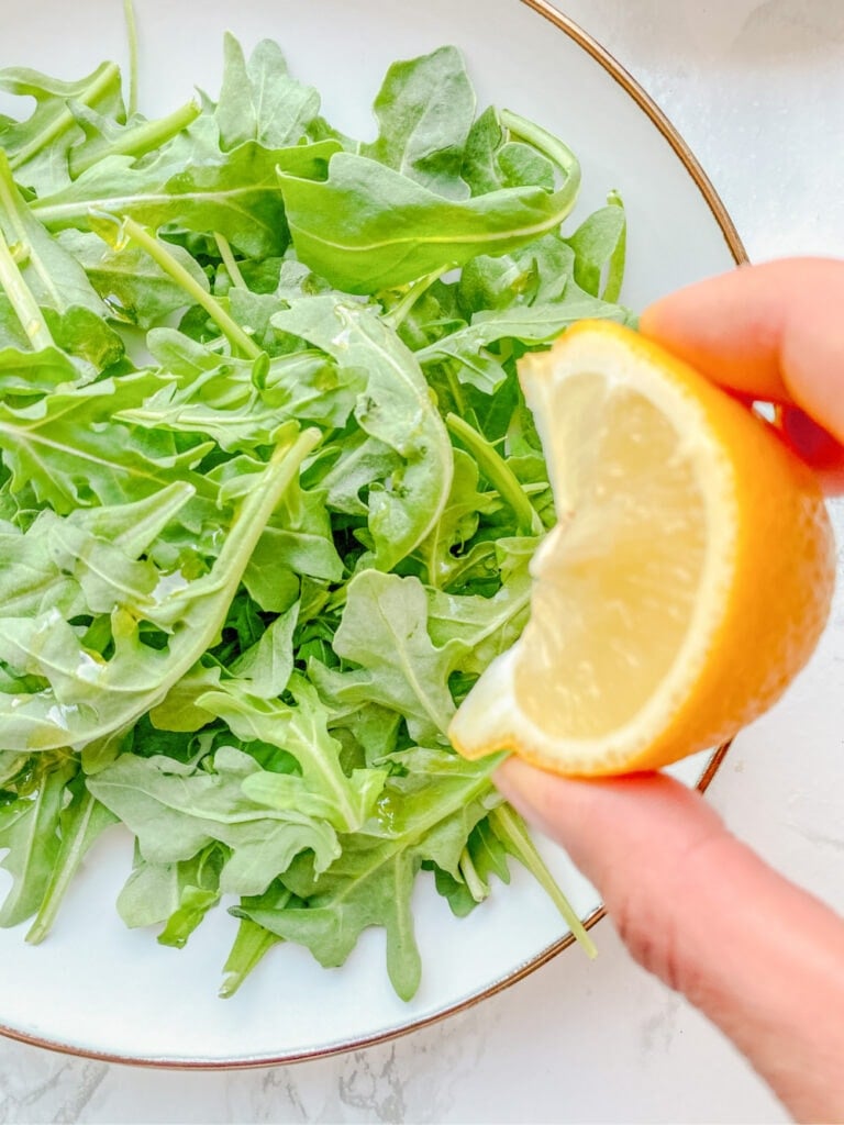 Close up photo of a hand squeezing a slice of lemon onto a bed of arugula on a white plate.