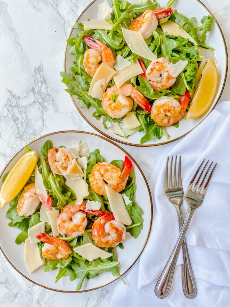 Overhead photo of two arugula salads topped with shrimp, shaved parmesan cheese, and lemon slices. There are two forks forming an X on a white napkin off to the side.