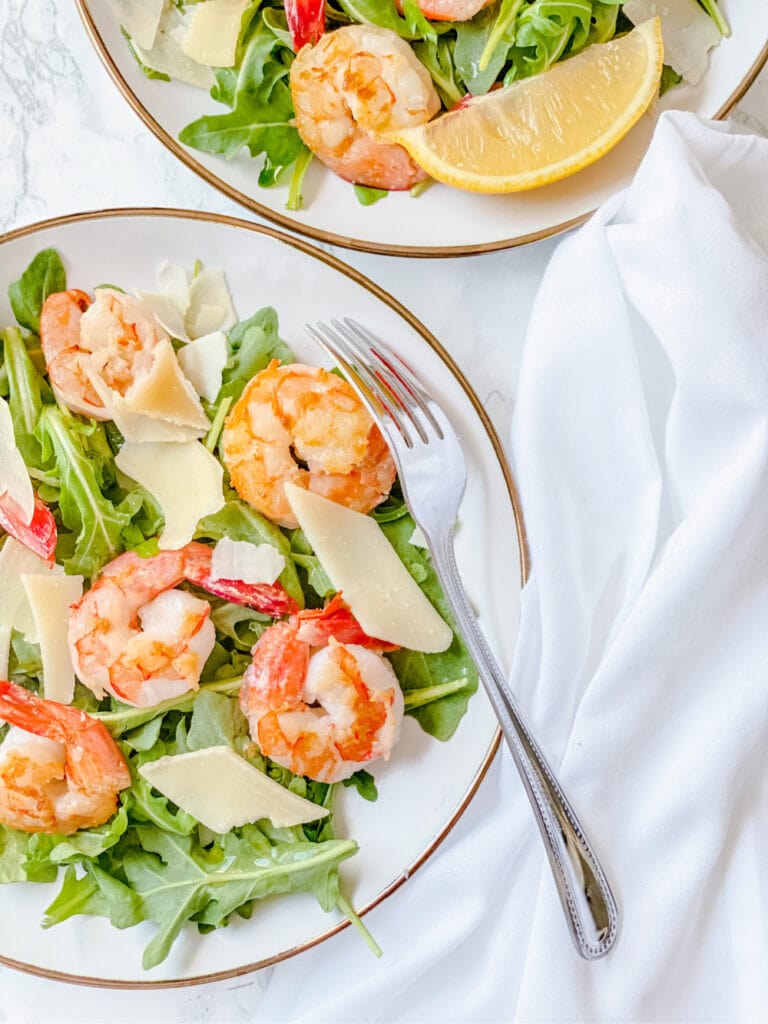 Two plates of arugula salad with shrimp and Parmesean on a white background with a fork and white napkin on the side.
