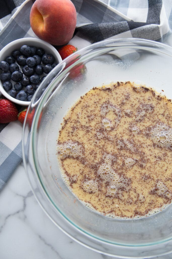 A glass bowl of french toast custard with cinnamon sitting next to a bowl of blueberries and a peach