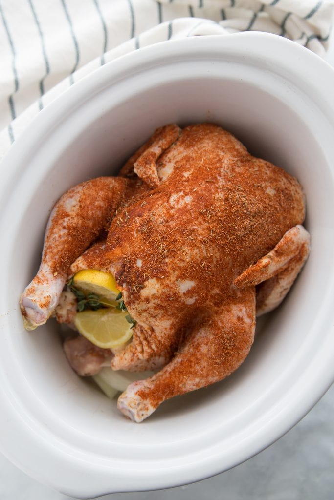 A whole chicken covered in spices sitting in a white ceramic crock pot insert
