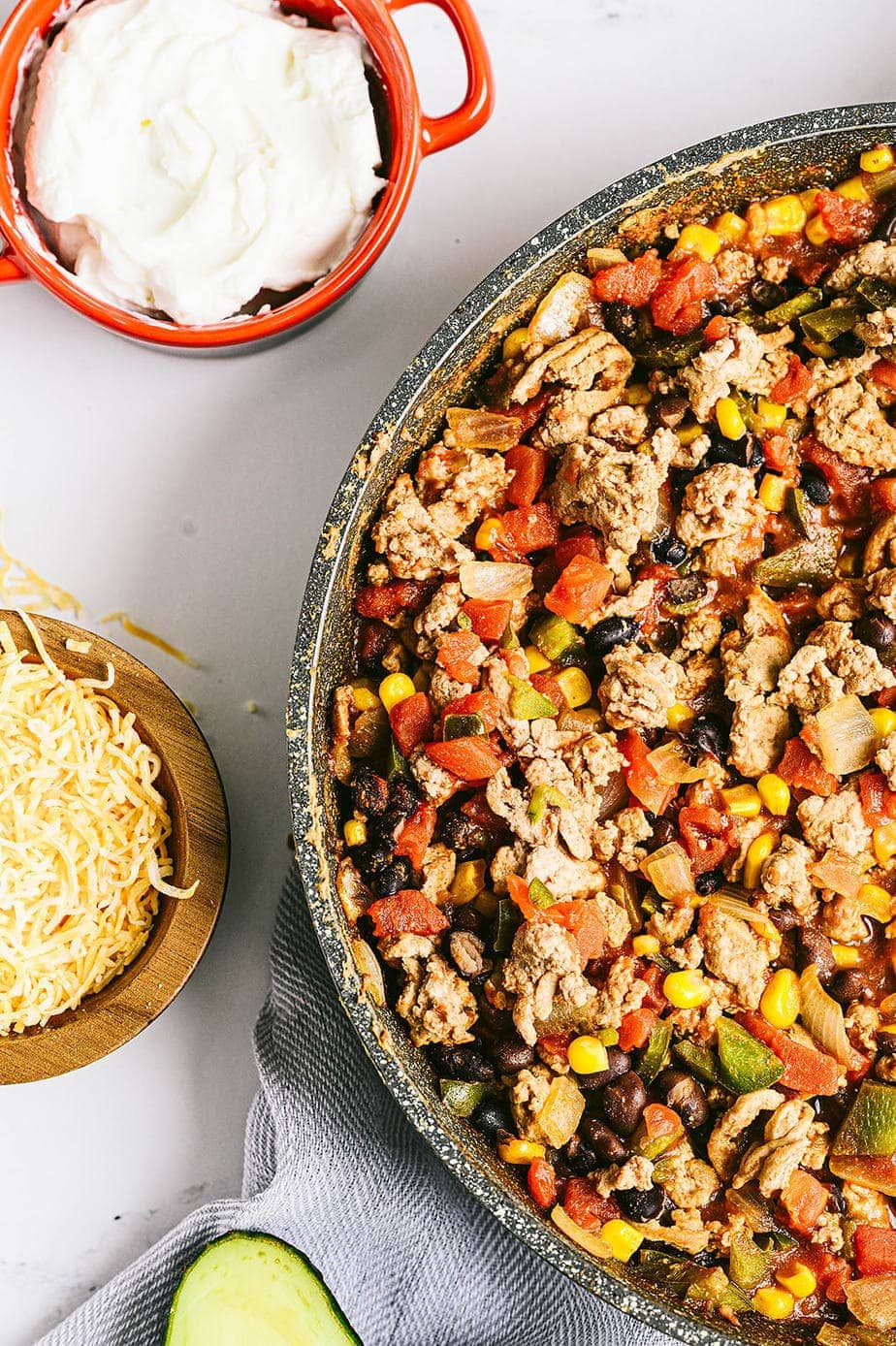 An overhead shot of a skillet with ground turkey, bell peppers, beans, corn, and onions. Yogurt, cheese, and avocado sit nearby as toppings