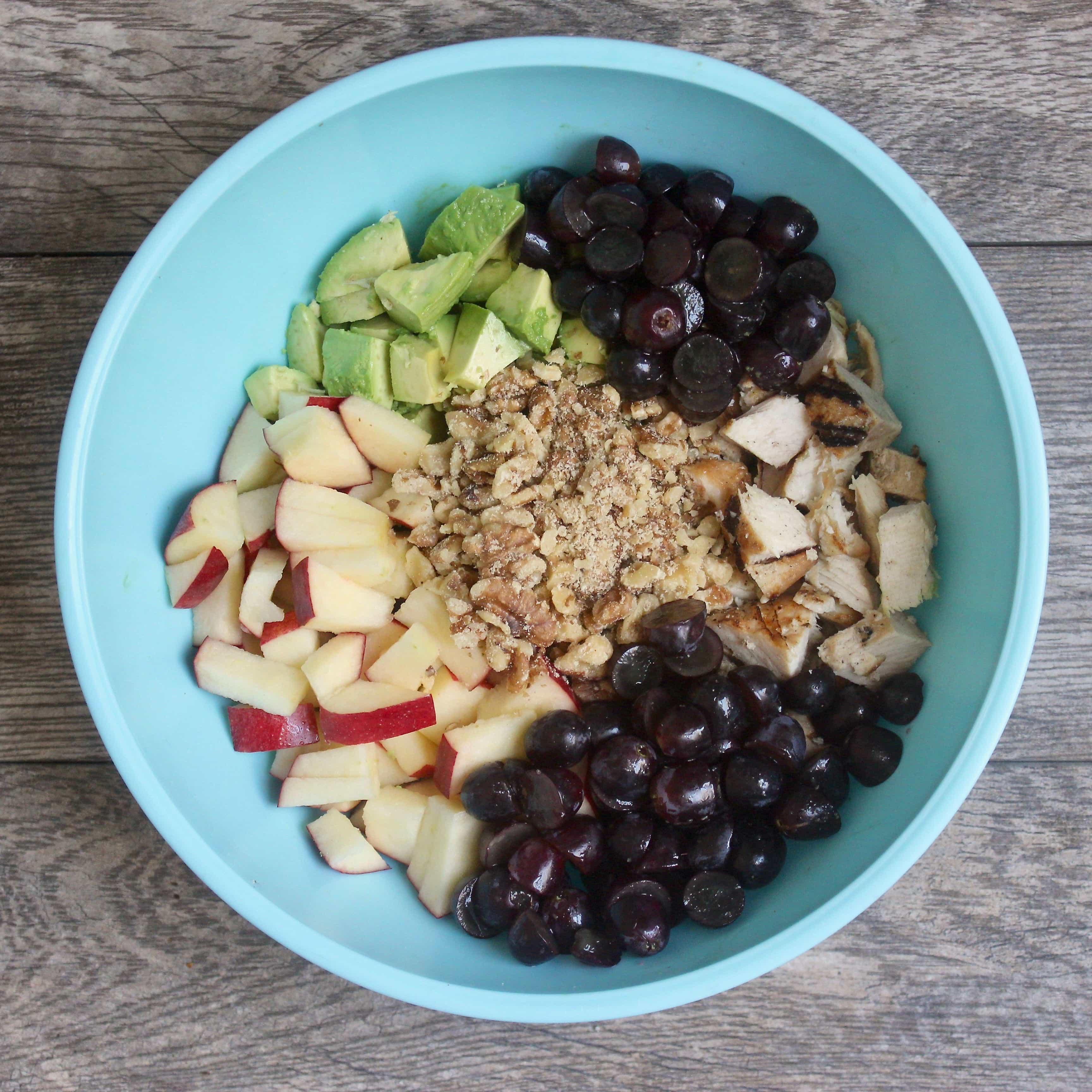 Diced chicken, sliced graps, diced walnuts, diced appples, and diced avocados sit in a  blue bowl, unmixed on a wooden surface