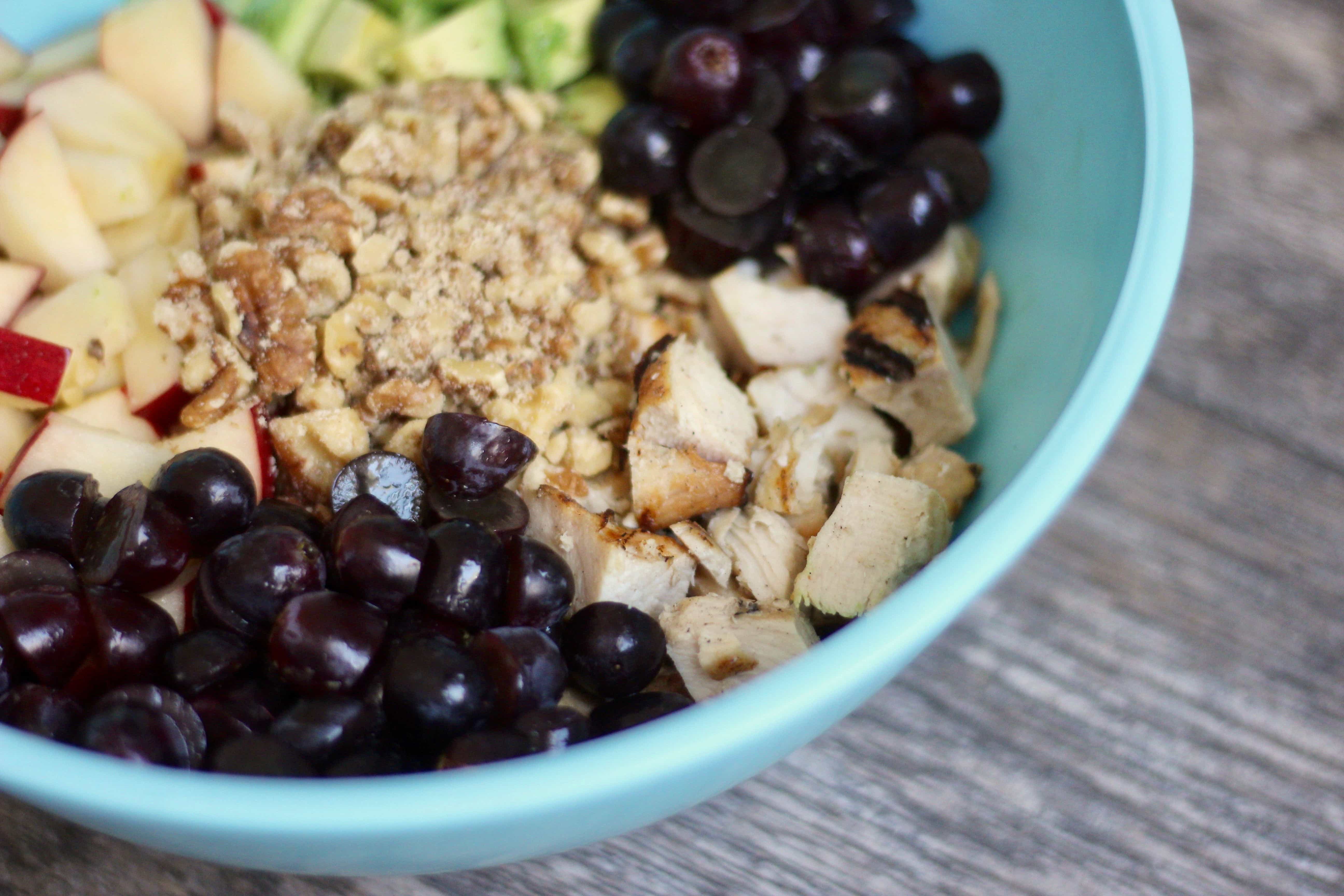 Diced chicken, sliced graps, diced walnuts, diced appples, and diced avocados sit in a  blue bowl, unmixed on a wooden surface