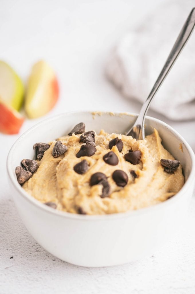 A bowl of healthy cookie dough with a spoon and several apples in the background