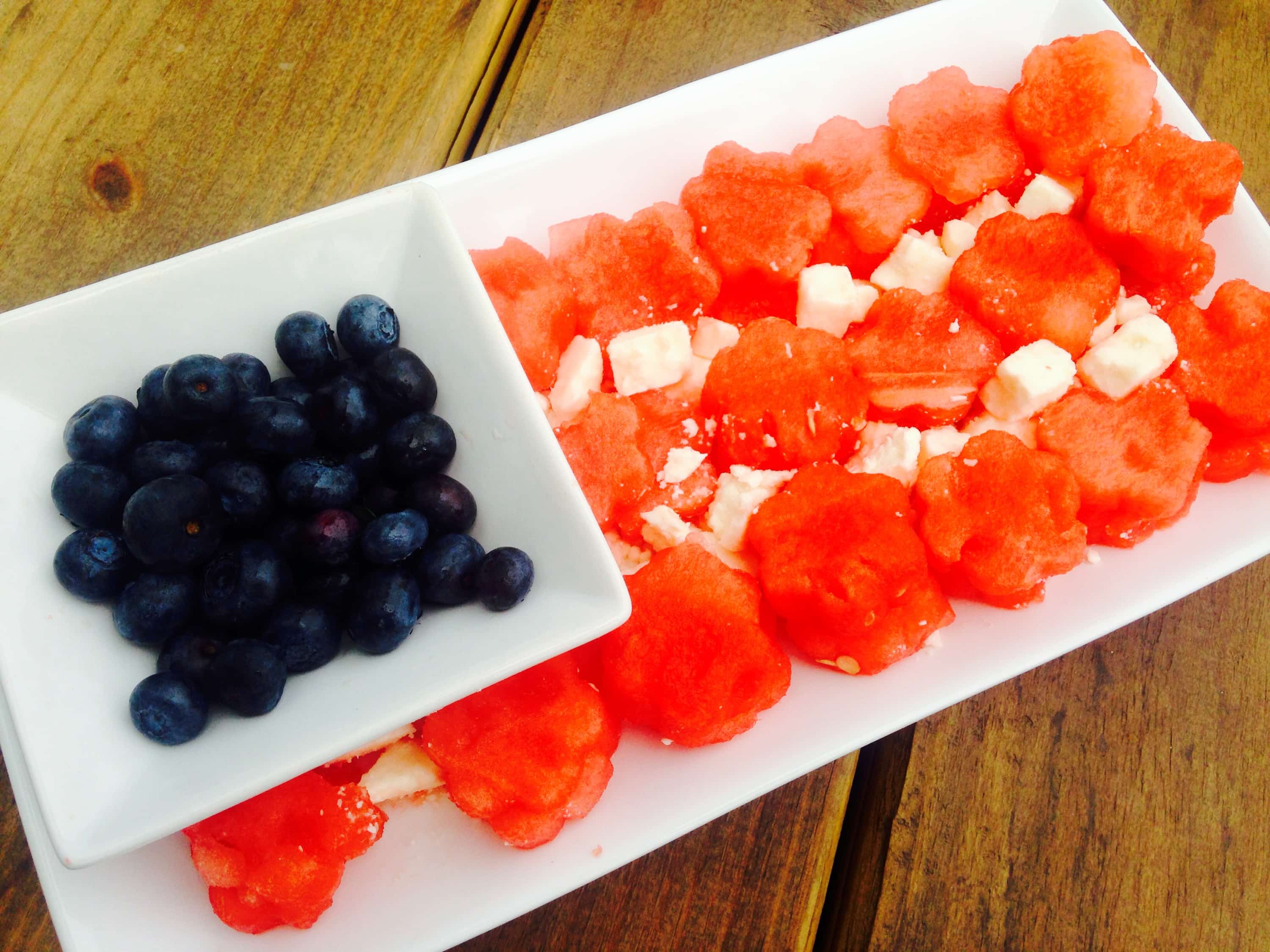 Watermelon, feta, and blueberries arranged in the shape of an American flag on a long rectangular serving plate with a wooden backgrouund