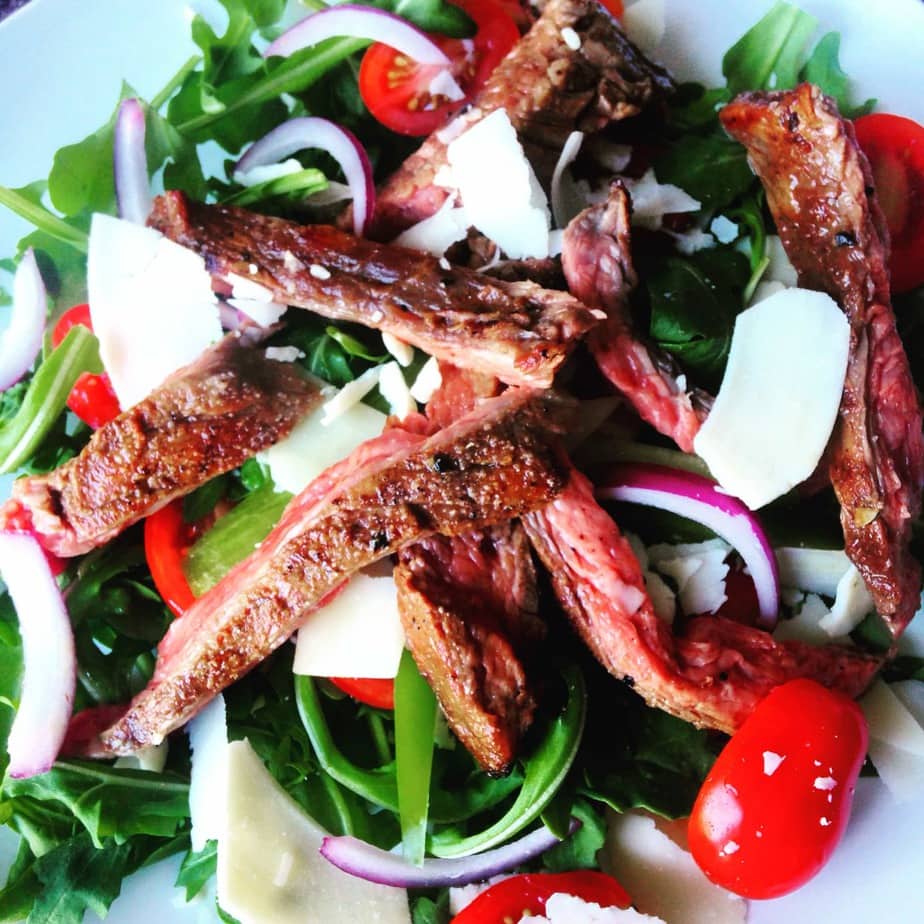 Fresh arugula topped with halved grape tomatoes, sliced red onions, shaved Parmesan cheese and Flank Steak cut into strips on a white plate