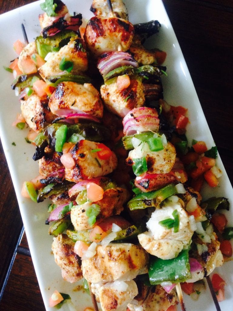 Grilled southwest chicken, green pepper, and red onion alternate on wooden skewers. Many are piled on a long rectangular plate with a dark background.