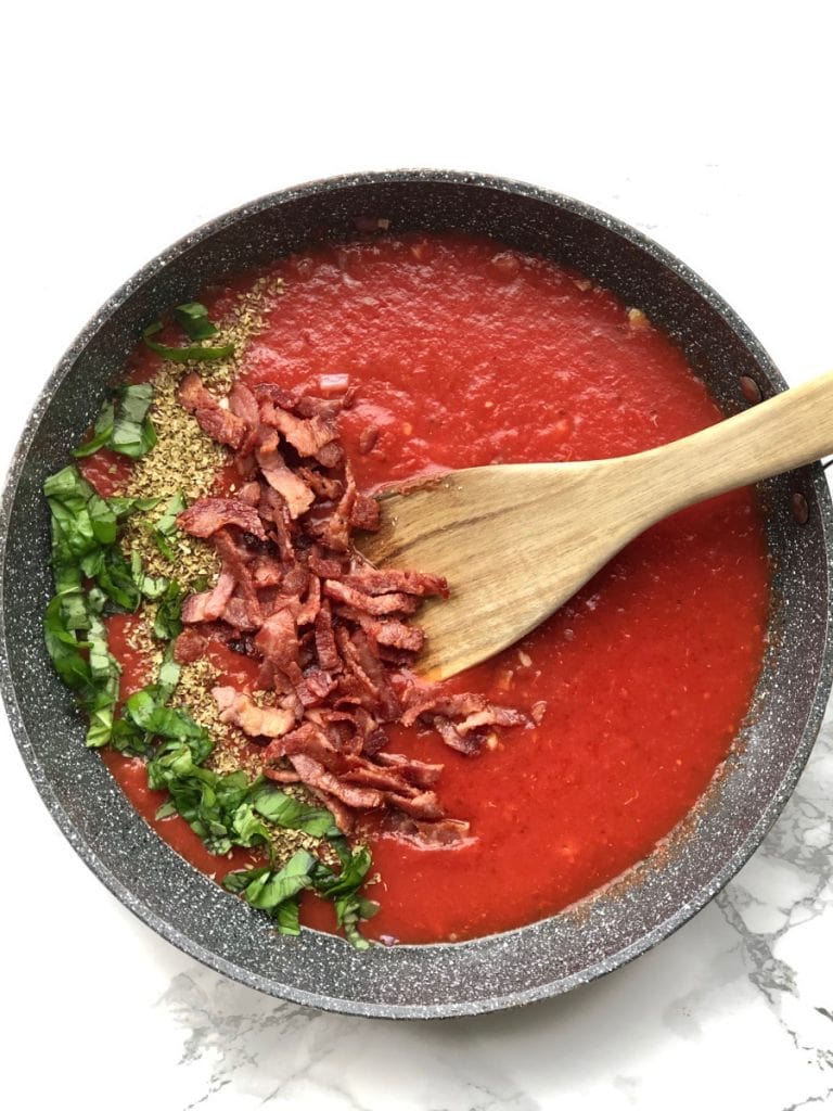 A overhead shot of a skillet with red sauce, basil, oregano, and turkey bacon. 