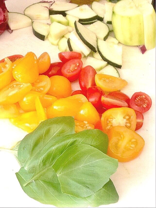 Fresh basil, halved tomatoes, and sliced zucchini are displayed on a cutting board