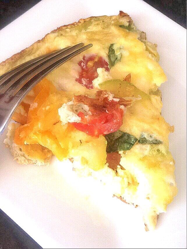 A triangular slice of fritatta on a white plate with tomatoes, zucchini, and basil peeking through the egg dish