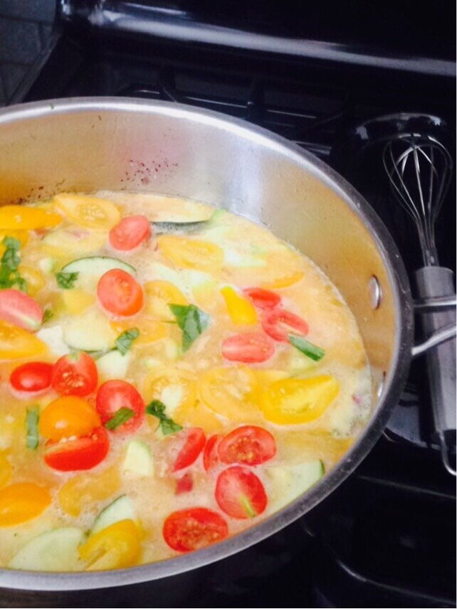 Red and yellow tomatoes, basil, and zucchini begin to cook in a pan with beaten eggs. A whisk lays to the side.