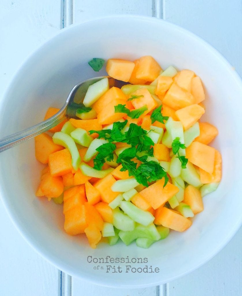 Diced cucumber and cantaloupe in a white bowl topped with fresh mint and a silver spoon resting on the side