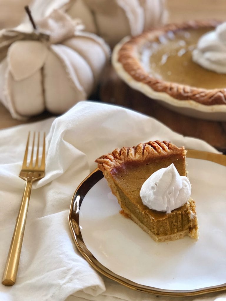 A piece of Healthy Pumpkin Pie is on a white plate rimmed with gold. Next to the plate is a gold fork on a white fabric napkin and the rest of the pumpkin pie is set in the background. The pie is topped with coconut cream. 