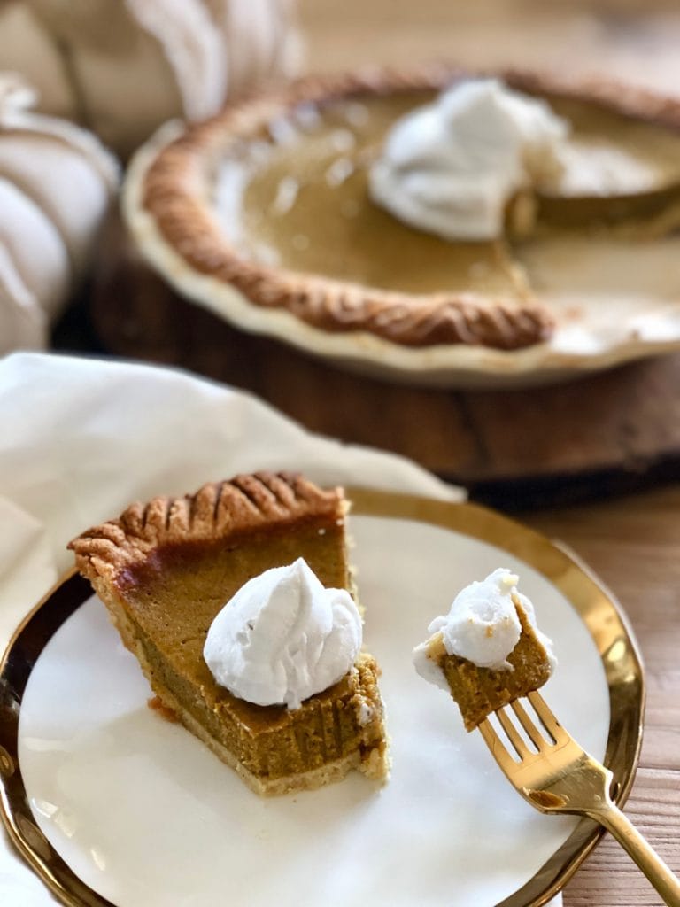 A piece of Healthy Pumpkin Pie is on a white plate rimmed with gold. A piece of pie is on a gold fork and the rest of the pumpkin pie is set in the background. The pie is topped with coconut cream. 