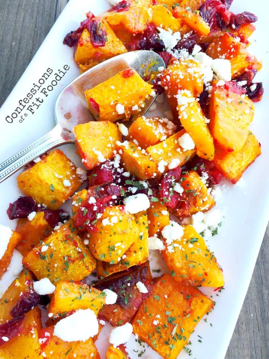 Cubed roasted butternut squash and roasted cranberries topped with crumbled goat cheese and parsley, on a white rectangular plate, on a wooden background. 