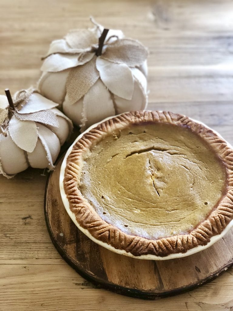 Baked Healthy pumpkin pie in a white pie plate on a wooden round, resting on a wooden table. There are two cream colored fabric pumpkins sitting next to the pie.