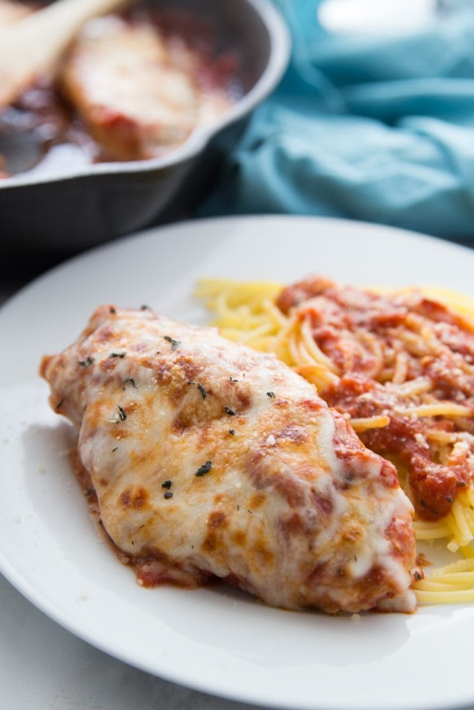 Chicken breast topped with tomato sauce and melted cheese next to spaghetti and tomato sauce on a white plate- the rest of the pan of chicken sits in the background