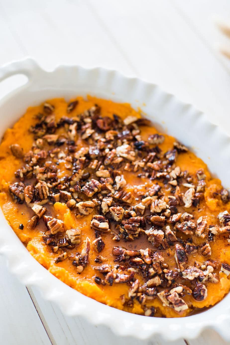Close up photo of healthy sweet potato casserole in a white oval baking dish on a white wooden background. The mashed sweet potatoes are topped with a toasted pecan streussel.