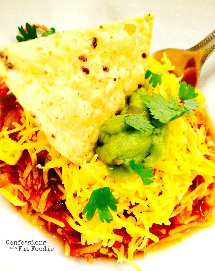 Crock Pot Chili Chicken topped with cheddar cheese, cilantro, avocado, and a single chip dipped in the middle. A fork is resting on the side of the bowl.