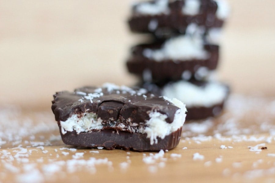 Close up side view of a homemade chocolate coconut candy cup with a bite taken out.  Out of focus in the background is a stack of 3 coconut cups.  There is coconut sprinkled on the wooden tabletop.