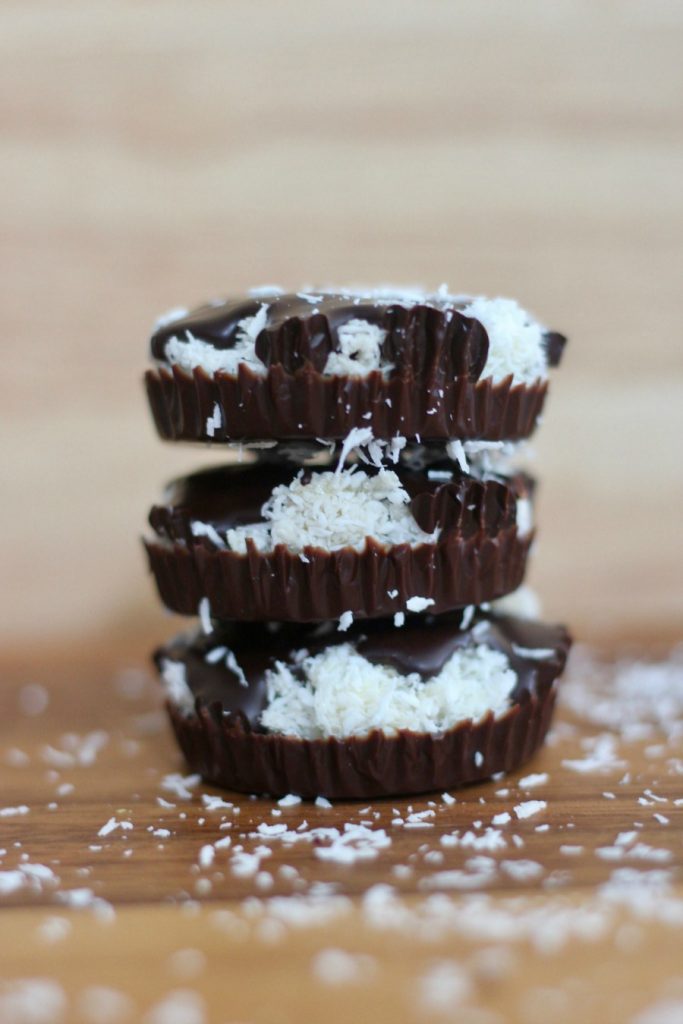 Close up side view of a stack of 3 homemade chocolate coconut cups- copycat mounds candy.  There is coconut sprinkled on the wooden tabletop.