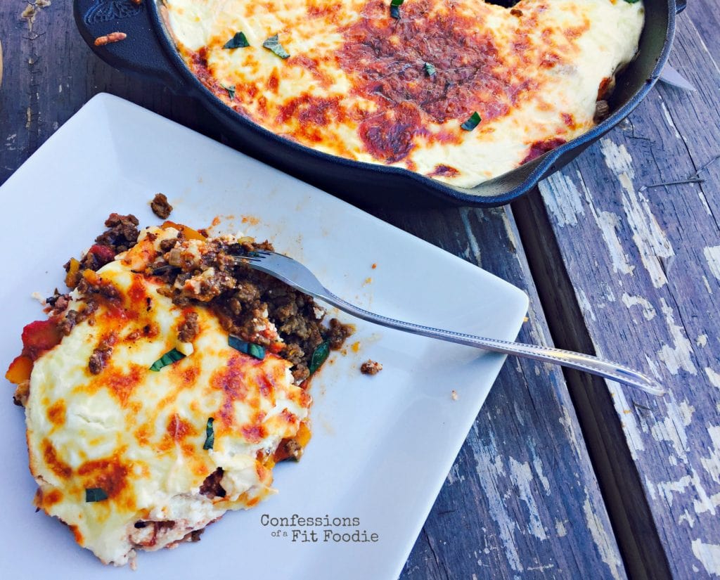 Photo of a serving of lazy butternut squash lasagna on a white plate with a fork ready to dig in. Ground beef and tomato sauce peek out from a layer of crispy lasagna cheese and garnished with some basil. In the background is the rest of the lasagna in a cast iron skillet sitting on a wooden table.