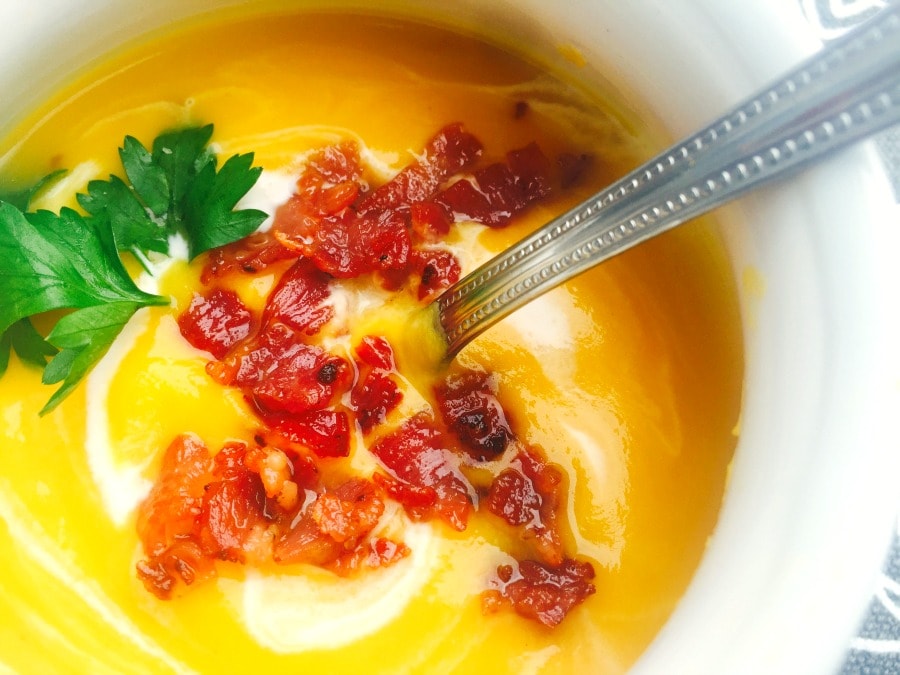 Close up photo of a white soup mug filled with dairy free, gluten free butternut squash soup, swirled with coconut milk and topped with crispy turkey bacon and fresh parsley, with a spoon in it.