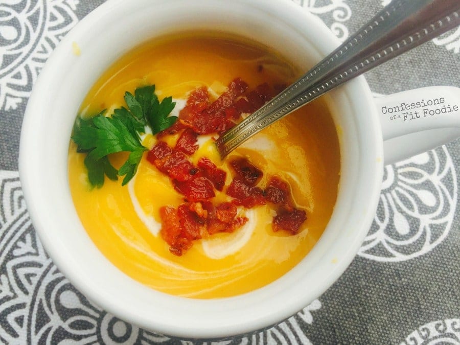 Overhead photo of a white soup mug filled with dairy free, gluten free butternut squash soup, swirled with coconut milk and topped with crispy turkey bacon and fresh parsley. The mug is sitting on a gray and white paisley tablecloth.
