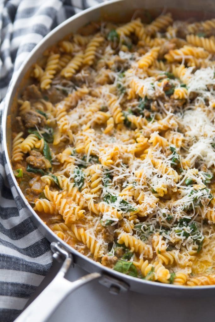 large saucepan with pumpkin pasta and spicy sausage topped with shredded parmesan cheese. pan is resting on a blue and white checkered napkin.