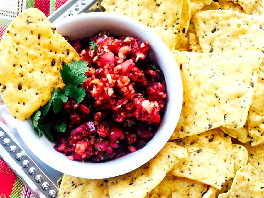 Close up photo of Holiday Salsa in a white bowl with tortilla chips on a silver platter. A festive green and red towel is in the background.