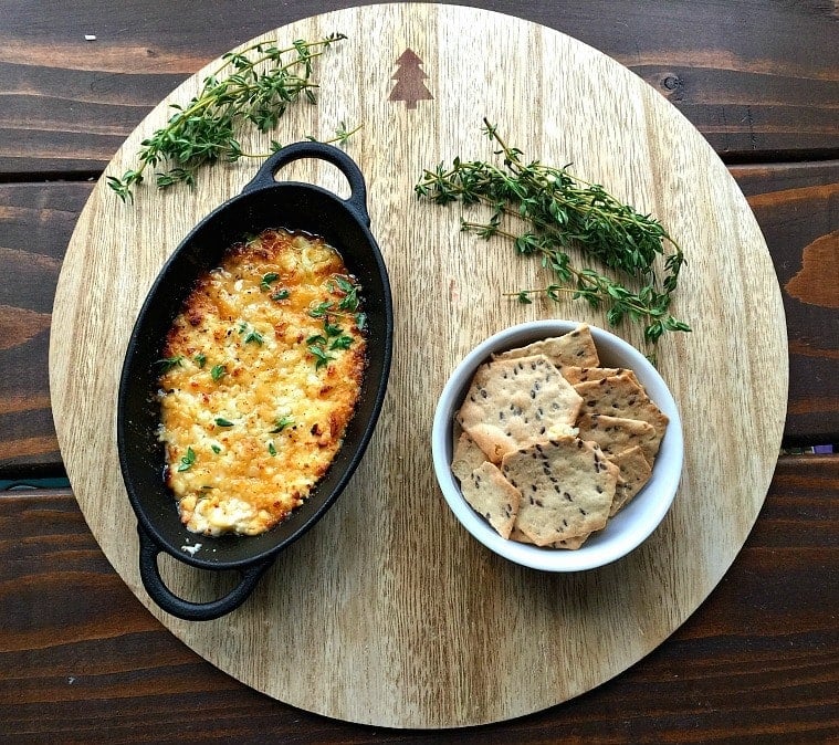 Cast iron pan of baked feta dip next to a white bowl of gluten free crackers on a wooden round, garnished with thyme.