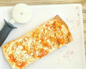 Personal flatbread pizza topped with buffalo chicken, ricotta, blue cheese, and mozzarella cheese laying diagonally on a cutting board next to a pizza cutter. Recipe from the blog, Confessions of a Fit Foodie.