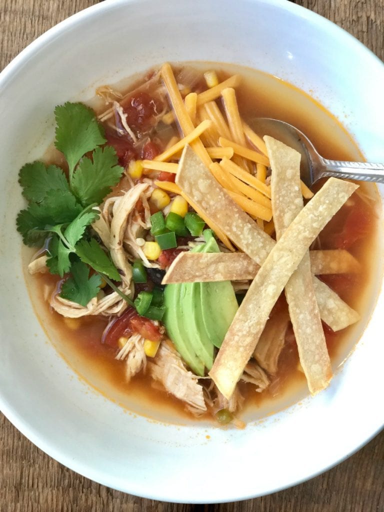 Overhead photo of chicken tortilla soup topped with shredded cheddar, avocado, tortilla strips, and cilantro.
