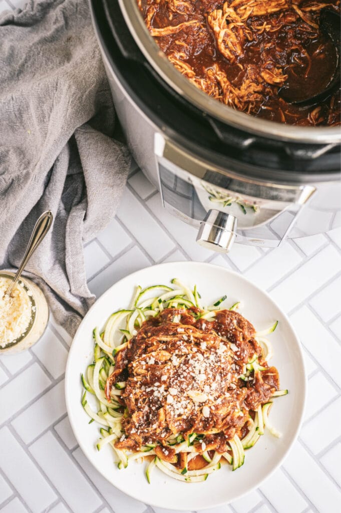 Overhead photo of chicken in the Instant Pot with a plate of chicken and zoodles on a white, tile counter top.