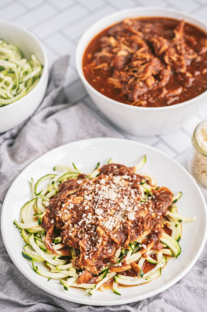 A white plate of balsamic chicken over spiralized zoodles and topped with parmesan cheese. In the background are two large bowls, one is full of plain zoodles, the other is full of balsamic chicken and sauce.