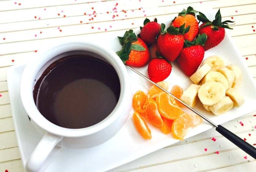  Una pequeña olla blanca de fondue de chocolate sin lácteos en un plato rectangular blanco. En el plato hay fresas enteras, plátanos en rodajas y segmentos de mandarina. Hay pequeños trozos de confeti en forma de corazón sobre el fondo de madera blanca.