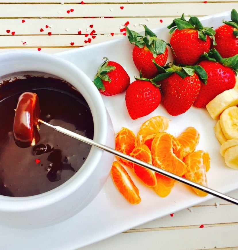 Olla de fondue blanca rellena de chocolate con un segmento de mandarina sumergido con un tenedor de fondue metálico. En el lado del plato rectangular hay fresas enteras, plátanos en rodajas y segmentos de mandarina. 