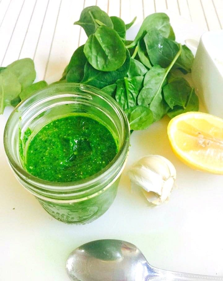 Side view of a mason jar full of homemade spinach pesto sauce on a white cutting board with a white wooden background too. Next to the jar are some ingredients to make it- a pile of spinach leaves, half of a lemon, half of a bulb of garlic, and an overturned spoon. 
