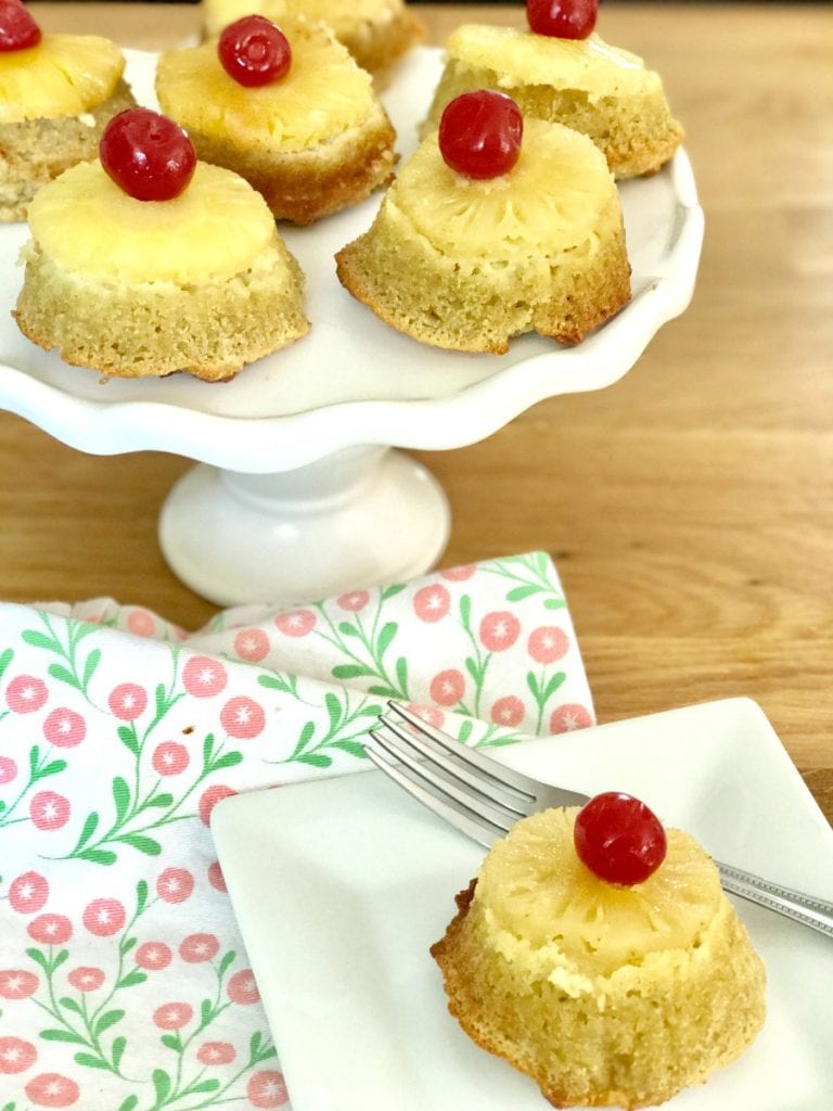 Cupcakes topped with pineapples and cherries on a white cake stand with one cupcake on a square plate with a fork.