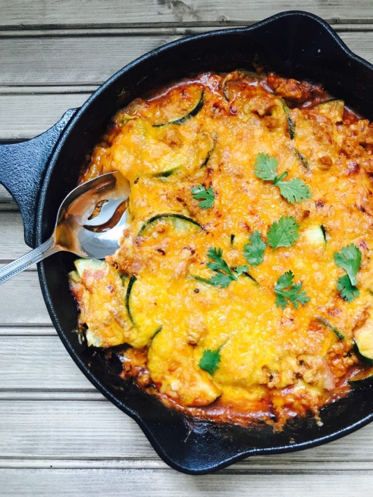 Cast iron skillet on wooden background filled with zucchini and ground meat, topped with cheese and cilantro. A serving spoon is poised on the side, ready to dig in.