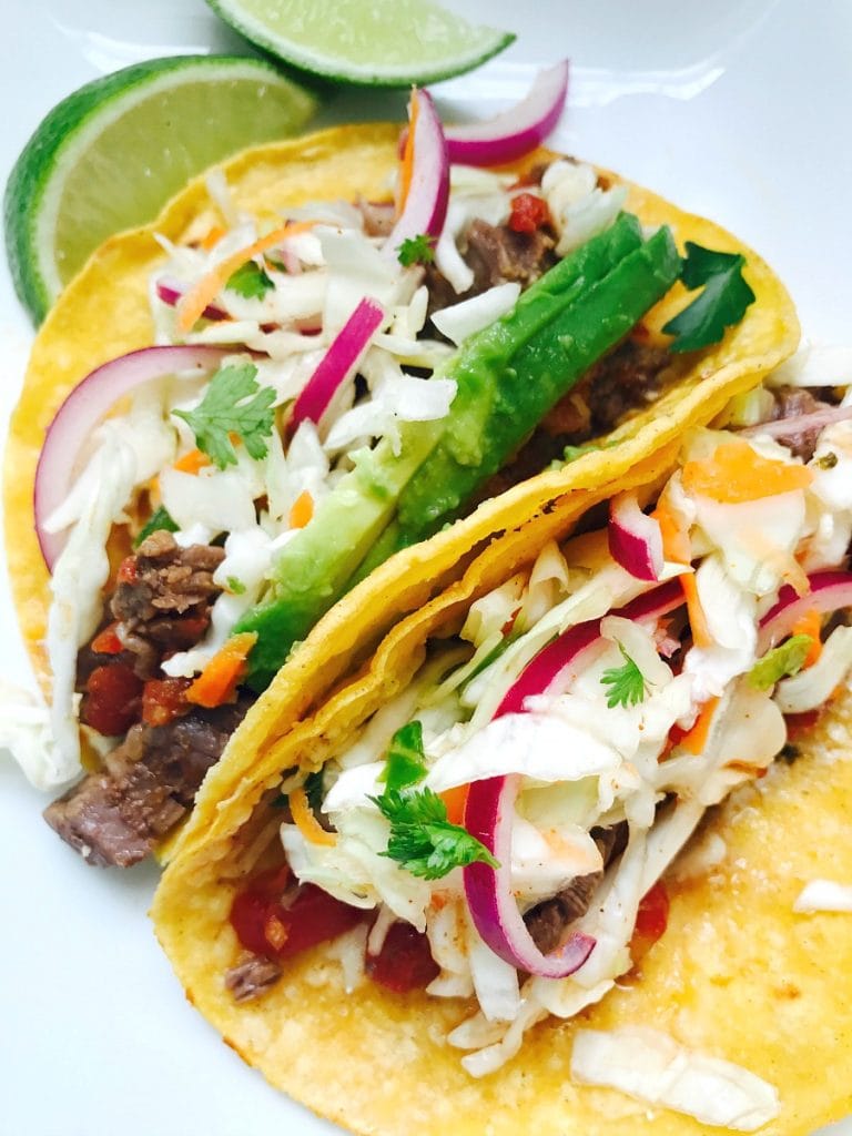 Two Instant Pot Flank Steak Tacos sit propped side by side on a white plate with limes in the background. In the tacos are flank steak, cilantro lime slaw, and topped with cilantro and avocado.