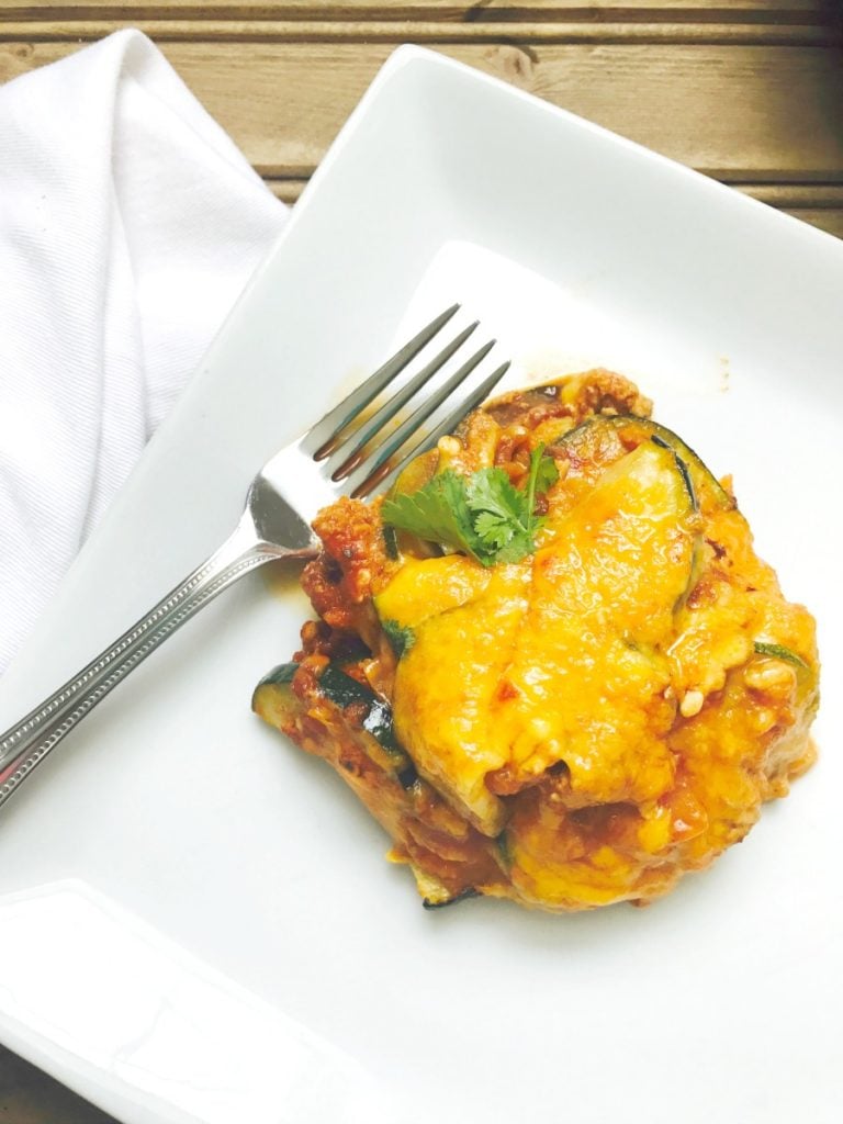 One neatly served portion of zucchini enchilada skillet on a square white plate and a fork on the side. Beneath the plate is a white napkin and wooden table.