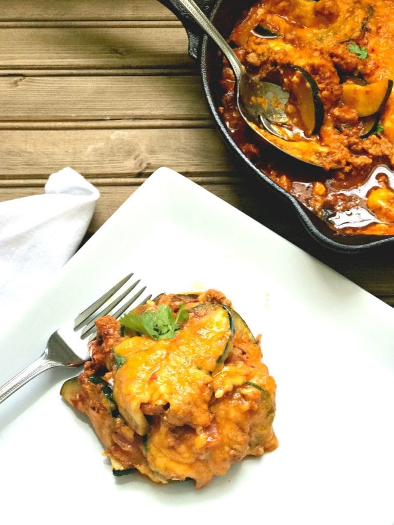 A portion of lazy zucchini enchilada skillet sits on a square white plate, a fork poised for eating. In the background is the rest of the cast iron skillet ready for serving. This all sits on a wooden table.