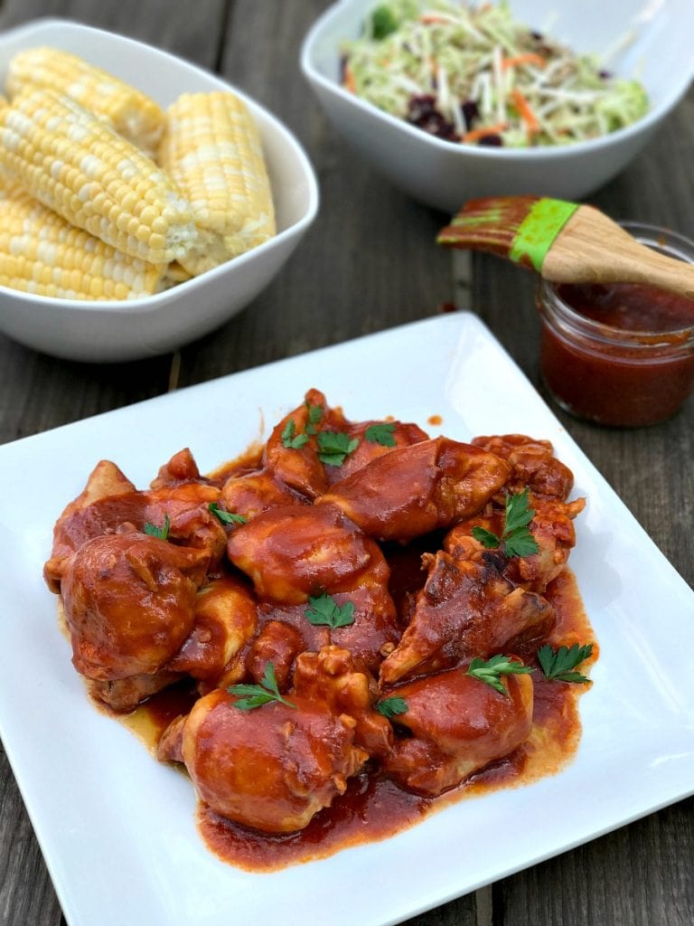 Maple chicken thighs in BBQ sauce on a square white plate.  BBQ sauce, corn on the cob, and broccoli slaw in dishes in the background.