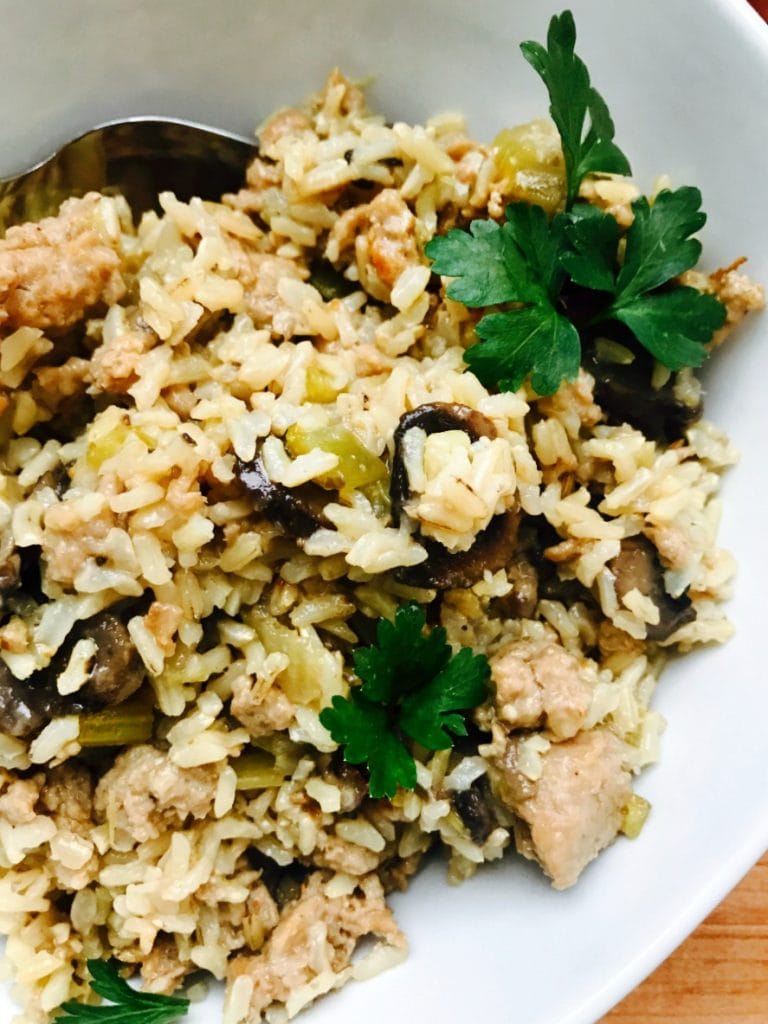 Close up photo of a white bowl full of Stuffing with brown rice, sausage, celery, and mushrooms and topped with fresh parsley.