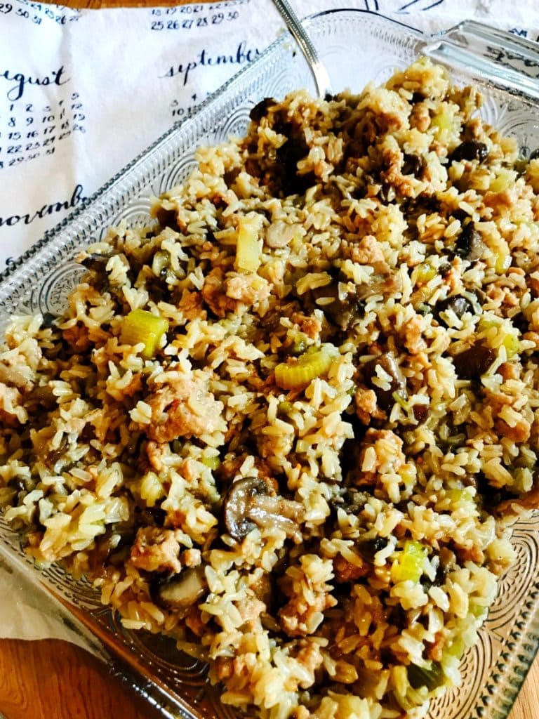 Rectangular glass baking dish full of Instant Pot Brown Rice Stuffing with a silver serving spoon on the side. In the background is a white and blue tea towel depicting a calendar.