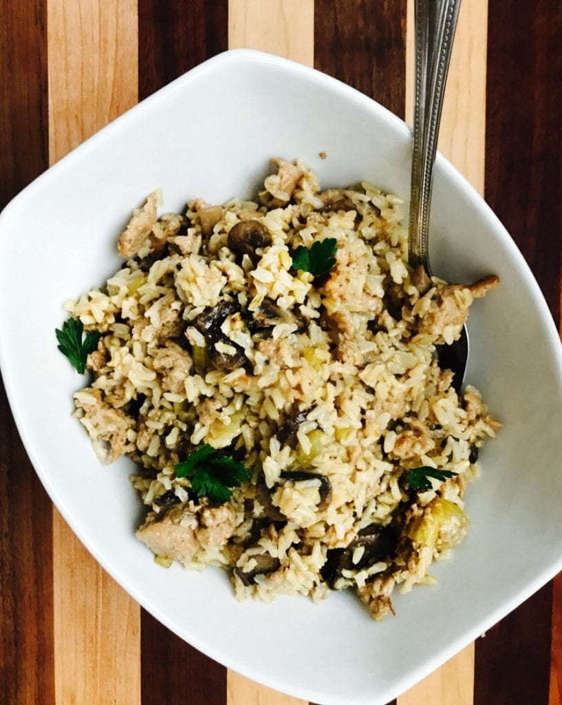 An overhead photo of a white rectangular bowl full of gluten free stuffing with sausage, packed with veggies like celery and mushrooms and topped with fresh parsley, on a striped wooden background.