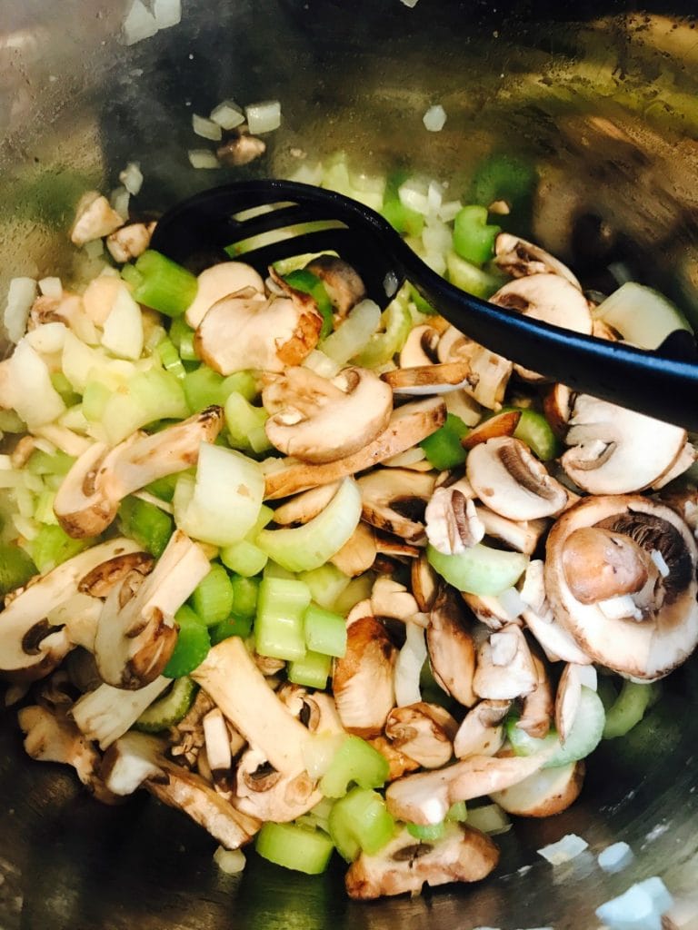 Raw celery, mushrooms, and onions sauteing in an Instant Pot with a black slotted spoon